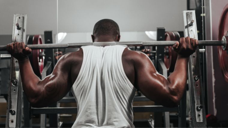 Man in white sleeveless t-shit squatting with a loaded barbell across his back