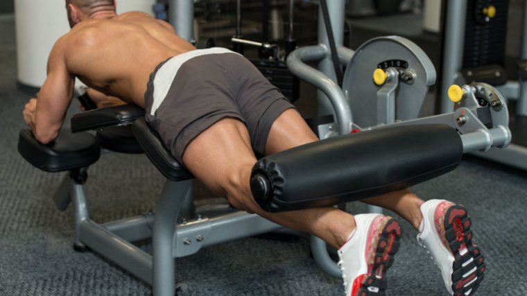 Person in gym doing lying hamstring curl on machine.